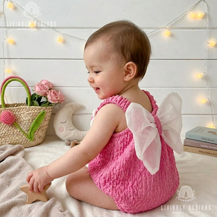 Side view of a baby sitting down wearing a bright pink textured romper with large white tulle angel wings attached to the back.