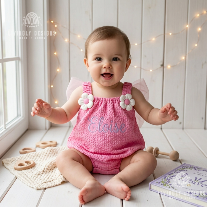 Smiling baby sitting on a white wooden floor wearing a pink textured romper with flower shoulder accents, tulle wings, and the name Eloise embroidered on the front.
