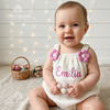 Smiling baby wearing a white textured romper with pink flower shoulder details and the name Emilia embroidered in pink.