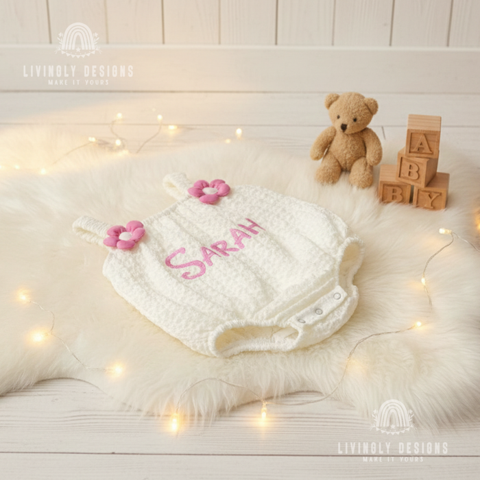 White textured knit baby romper laid flat on a white faux fur rug featuring pink flower shoulder details and the name Sarah embroidered in pink.