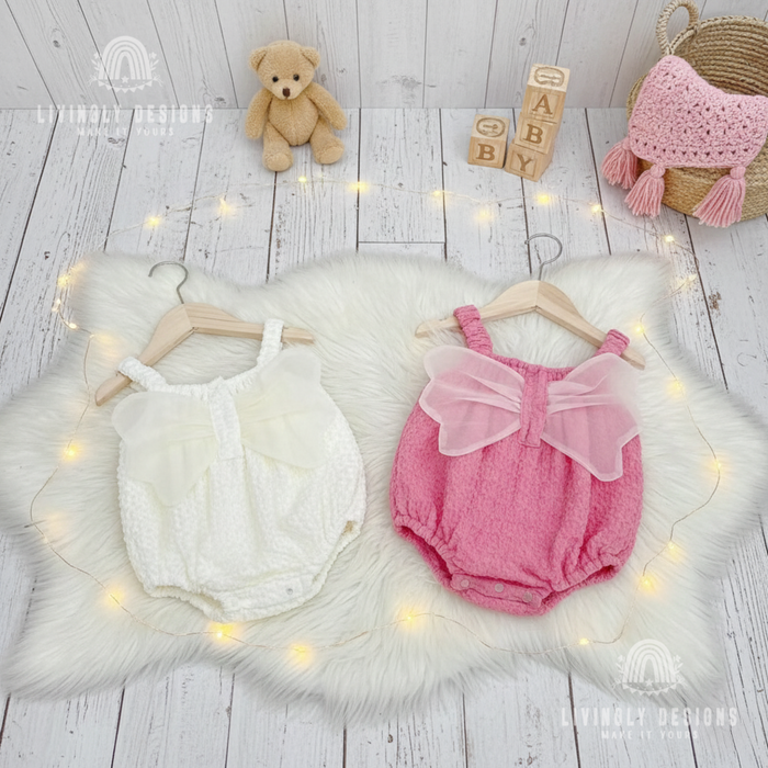 White and pink textured baby rompers with tulle angel wings displayed side by side on wooden hangers over a white faux fur mat.