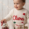 Smiling child wearing a cream knitted Christmas sweater personalized with the name “Clara” and festive Santa, Christmas tree, and holiday designs.