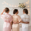 Three women wearing personalized satin bridal robes labeled Bride, Maid of Honor, and Bridesmaid.