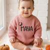 Smiling baby wearing a pink knitted sweater personalized with the name "Maya" in black lettering and an embroidered smiling crescent moon design.