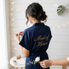 Woman wearing a navy satin robe embroidered with “Maid of Honor” on the back.
