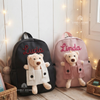 Black and pink backpacks personalized with handmade lettering spelling "Lucia" and "Linda," each featuring a plush teddy bear tucked into a front corduroy pocket