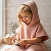 Young child wearing a dusty pink hooded waffle poncho bathrobe sitting near a window playing with a toy boat.