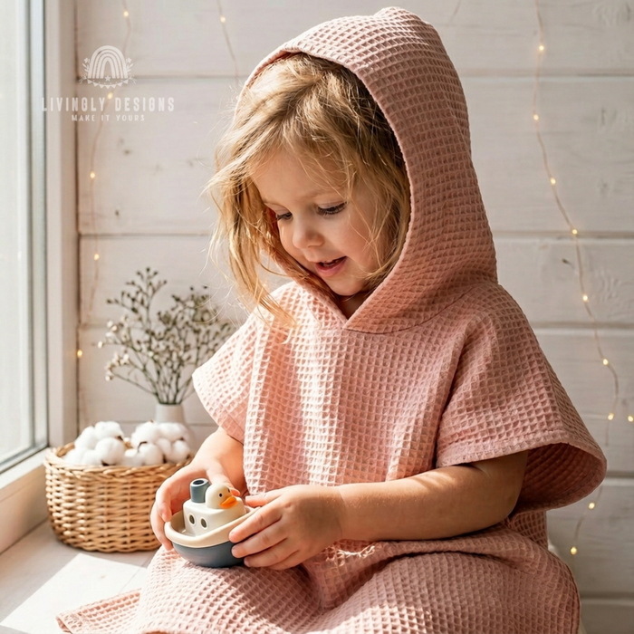 Young child wearing a dusty pink hooded waffle poncho bathrobe sitting near a window playing with a toy boat.