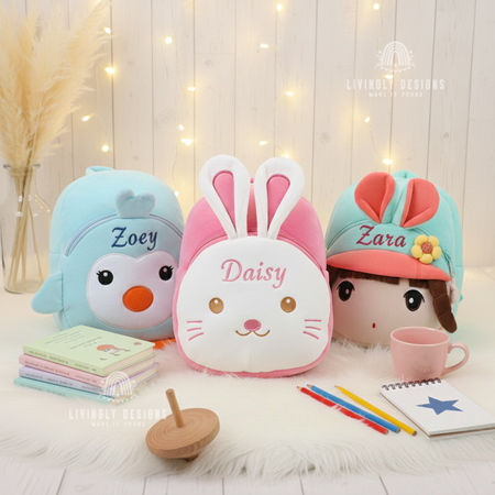 Three plush character backpacks displayed together on a white fur mat a blue penguin embroidered with Zoey, a pink bunny embroidered with Daisy, and a doll character with a cap embroidered with Zara surrounded by books, pencils, and a coffee mug.