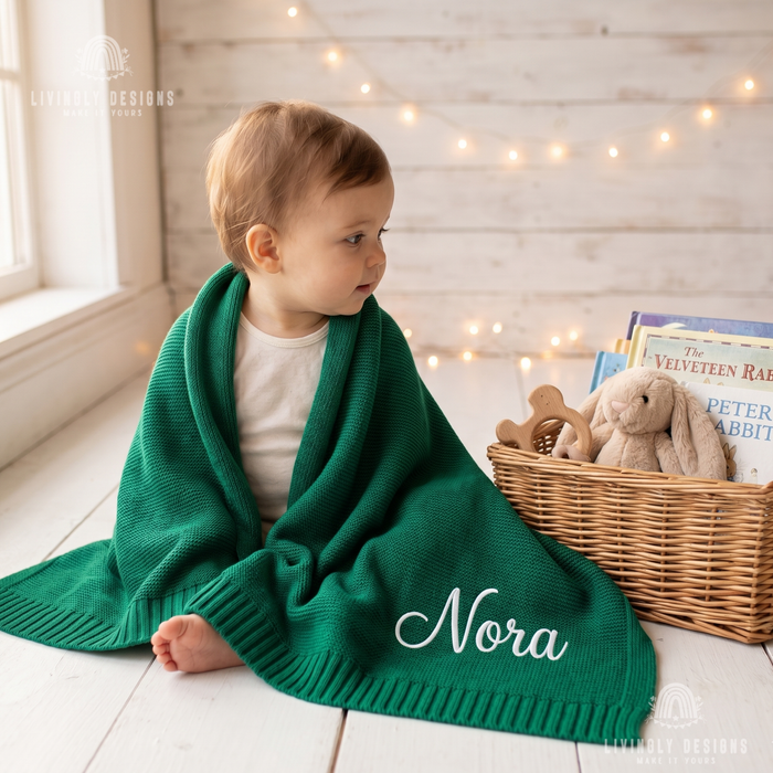 Baby wrapped in an emerald green personalized blanket named Nora, sitting next to a woven basket filled with books and a plush bunny.