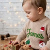 Child wearing a beige knitted Christmas sweater personalized with the name “James” and festive Santa, Christmas tree, and reindeer designs.