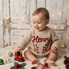 Smiling baby wearing a beige knitted Christmas sweater personalized with the name “Henry” and a reindeer design.
