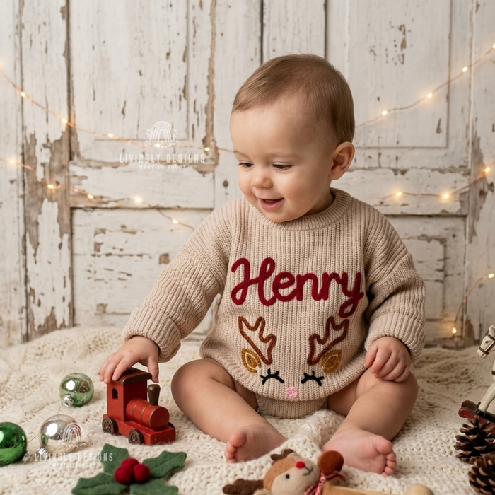 Smiling baby wearing a beige knitted Christmas sweater personalized with the name “Henry” and a reindeer design.