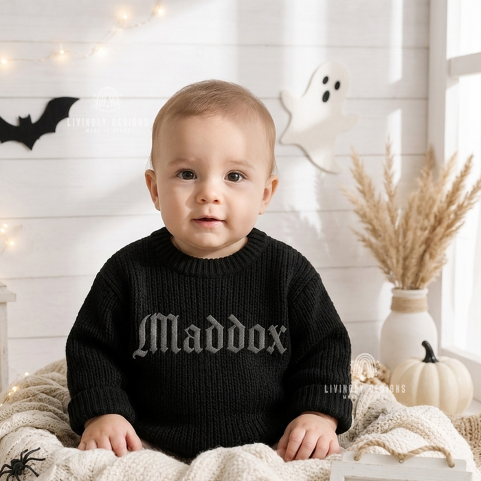 Baby wearing a black knitted sweater personalized with the name "Maddox," sitting near a ghost decoration and pampas grass.