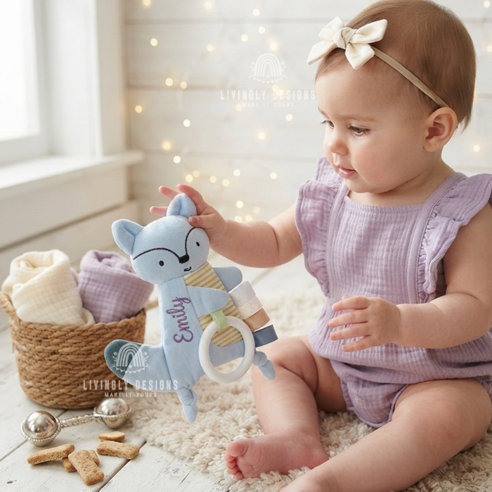 Baby girl in a purple outfit holding a light blue fox-shaped comforter with a teething ring, personalized with the name "Emily"