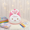 Plush pink bunny backpack with white bunny face, plush ears, and the name Clara embroidered on the forehead, displayed with books and colored pencils on a white fur mat.