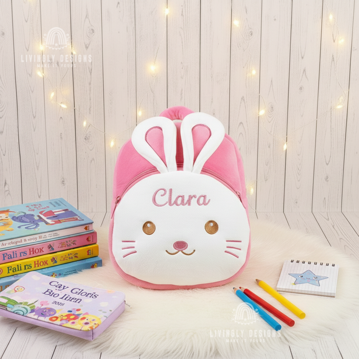Plush pink bunny backpack with white bunny face, plush ears, and the name Clara embroidered on the forehead, displayed with books and colored pencils on a white fur mat.