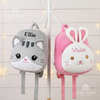 Plush grey kitten backpack embroidered with the name Ellie and plush pink bunny backpack embroidered with the name Violet hanging side by side on a white coat rack.
