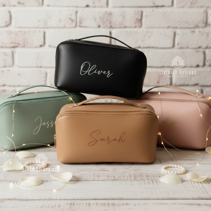 Four personalized leather cosmetic travel bags arranged against a white brick wall featuring custom cursive names—Oliver on black, Sarah on tan, Jessie on sage green, and Emily on light pink—surrounded by fairy lights and pearl jewelry.