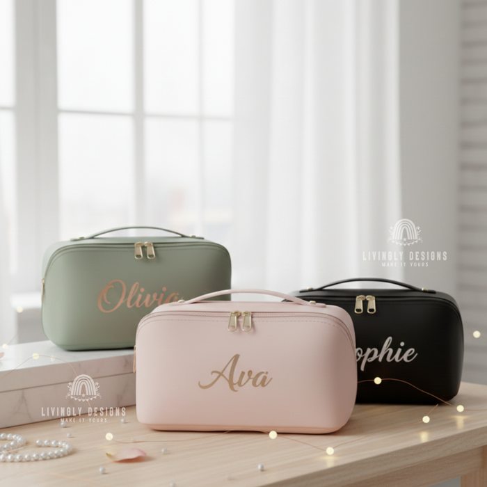 Three personalized leather cosmetic travel bags on a light wooden table—a sage green bag with Olivia, a light pink bag with Ava, and a black bag with Sophie—styled with pearl accessories and fairy lights in front of a window.