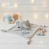 Plush grey elephant security blanket with knotted corners and the name Liam embroidered in blue, laying flat on a white wooden surface next to wooden baby teething toys and a blue plush bunny rattle.