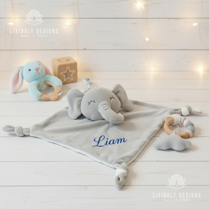 Plush grey elephant security blanket with knotted corners and the name Liam embroidered in blue, laying flat on a white wooden surface next to wooden baby teething toys and a blue plush bunny rattle.