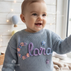 Smiling baby wearing a blue-grey knit sweater personalized with the name "Clara" in floral embroidery.