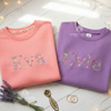Coral and purple sweatshirts personalized with the names "Eva" and "Evie" in colorful floral lettering, folded on a table