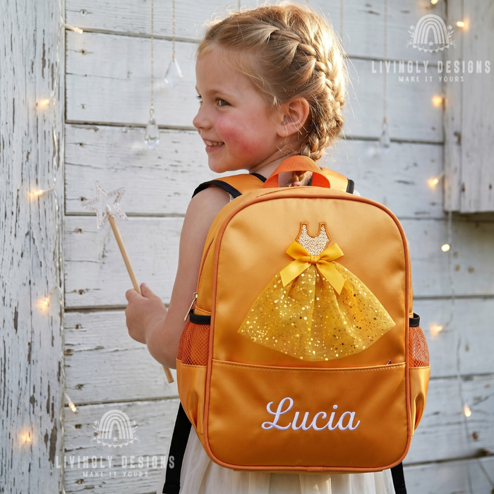 A young girl with blonde braided hair is shown from behind wearing a custom orange and gold backpack with a glittery dress design and the name 'Lucia' printed on the front. She is standing against a rustic white wooden wall with warm fairy lights.