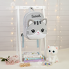 Plush grey kitten backpack embroidered with the name Sarah hanging on a white wooden display stand, accompanied by books, wooden blocks, and a white cat plush toy.