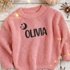Close up of a pink knitted sweater personalized with the name "Olivia" in black lettering, embroidered with a crescent moon and stars design.