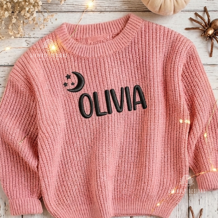Close up of a pink knitted sweater personalized with the name "Olivia" in black lettering, embroidered with a crescent moon and stars design.