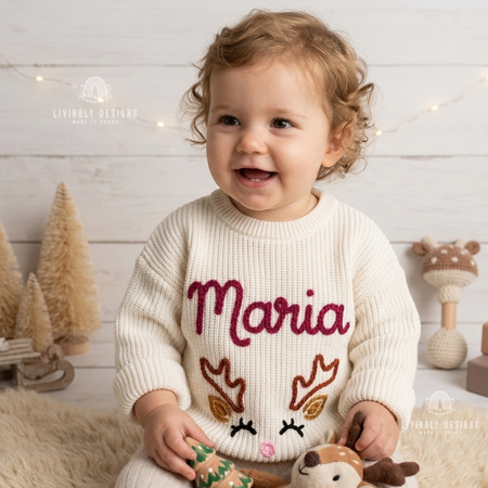 Smiling child wearing a cream knitted Christmas sweater personalized with the name “Maria” and a festive reindeer design.