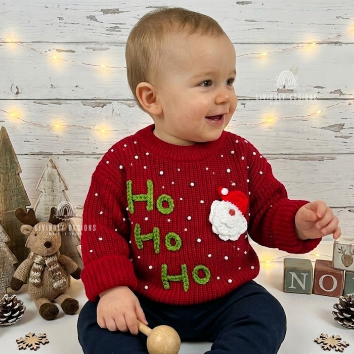 Smiling baby wearing a red knitted Christmas sweater with “Ho Ho Ho” lettering, white snow dots, and a Santa Claus design.