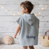 Back view of a young boy wearing a light blue hooded waffle poncho bathrobe custom embroidered with the name Henry in navy thread.
