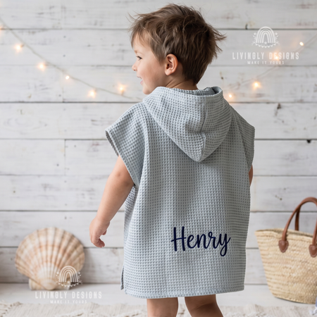 Back view of a young boy wearing a light blue hooded waffle poncho bathrobe custom embroidered with the name Henry in navy thread.