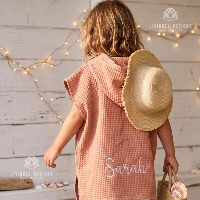 Back view of a young child wearing a dusty rose hooded waffle bathrobe custom embroidered with the name Sarah, carrying a straw bag and straw hat.