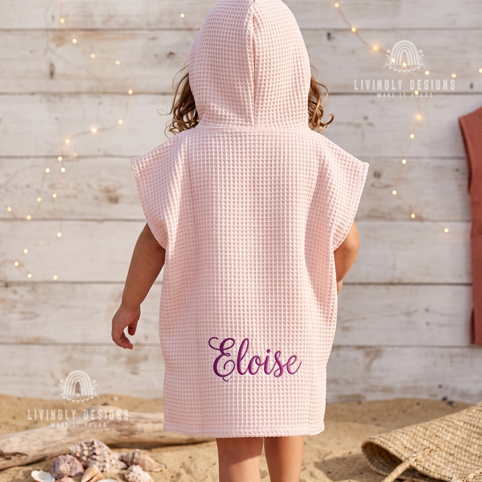 Back view of a toddler wearing a light pink hooded waffle poncho bathrobe custom embroidered with the name Eloise.