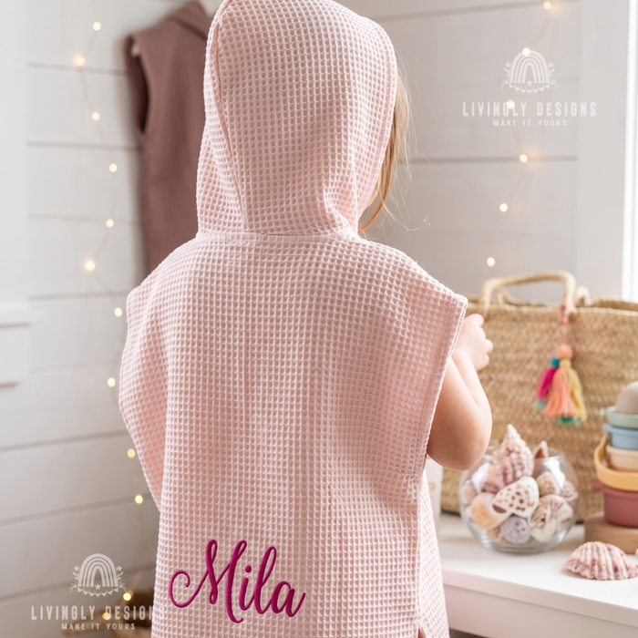 Back view of a young child wearing a light pink hooded waffle poncho bathrobe custom embroidered with the name Mila in magenta script thread.