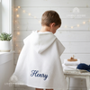 Back view of a young boy wearing a white hooded waffle poncho bathrobe custom embroidered with the name Henry in dark blue thread.