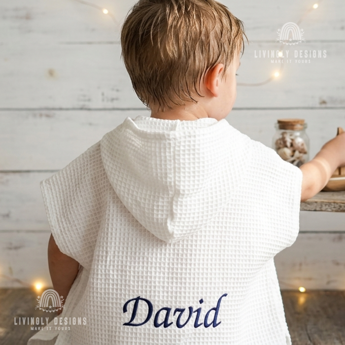 Back view close up of a young boy wearing a white hooded waffle poncho bathrobe custom embroidered with the name David in navy blue script thread.