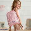 Smiling girl carrying a light pink backpack with a sparkly tulle ballerina dress appliqué, personalized with the name "Lucy," holding pink ballet shoes.