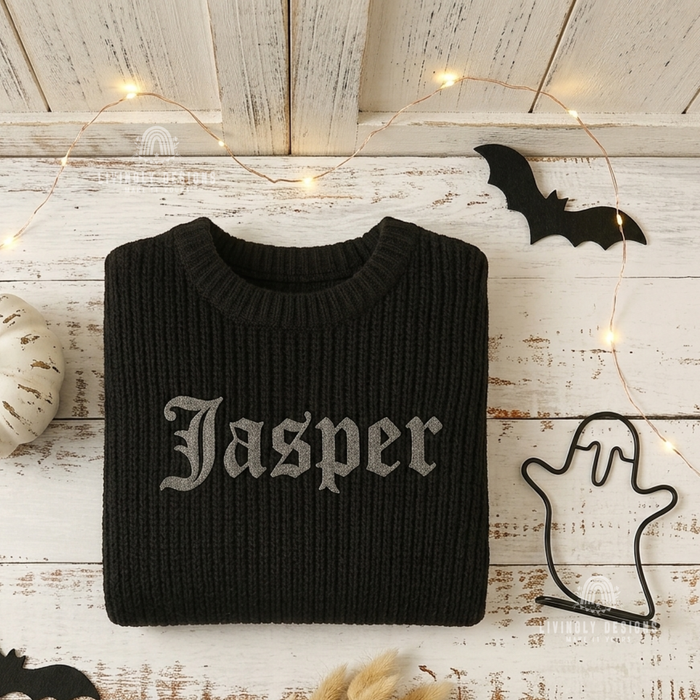 Folded black knitted sweater personalized with the name "Jasper" in gray gothic-style lettering