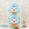 Two plush light blue penguin backpacks displayed on a white two-tier shelf unit the top embroidered with Olivia and the bottom embroidered with Arya accompanied by colored pencils and a small flower basket on a wooden background.