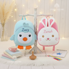Plush light blue penguin backpack embroidered with the name Zoey and plush pink bunny backpack embroidered with the name Zarah hanging side by side on a white wooden display stand, set on a white fur mat with books and pencils.
