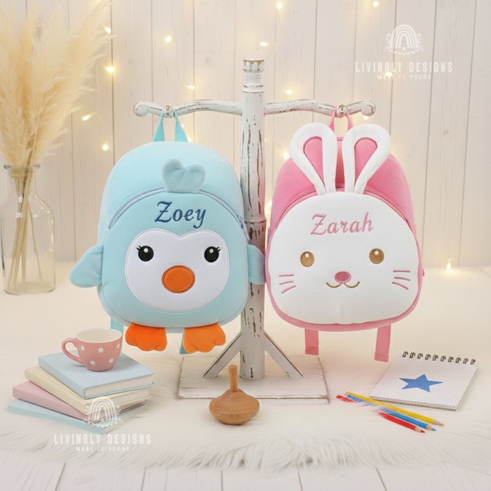 Plush light blue penguin backpack embroidered with the name Zoey and plush pink bunny backpack embroidered with the name Zarah hanging side by side on a white wooden display stand, set on a white fur mat with books and pencils.