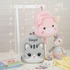Personalized plush animal backpacks displayed on a white stand a pink pig embroidered with Hailey hanging above and a grey kitten embroidered with Hazel sitting below, accompanied by a knitted rainbow unicorn plush toy, books, and crayons.