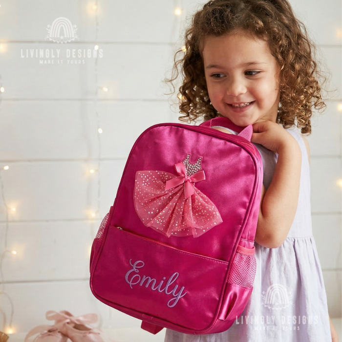 Smiling girl carrying a pink backpack with a sparkly tulle ballerina dress appliqué, personalized with the name "Emily"