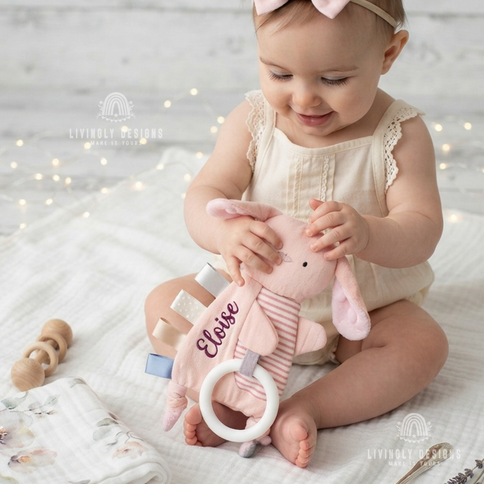 Smiling baby girl holding a pink bunny-shaped comforter with a teething ring, personalized with the name "Eloise"
