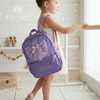 Girl wearing a cream tutu dress carrying a purple backpack with a sparkly tulle ballerina dress appliqué, personalized with the name "Luna," near a shelf with pink ballet shoes.